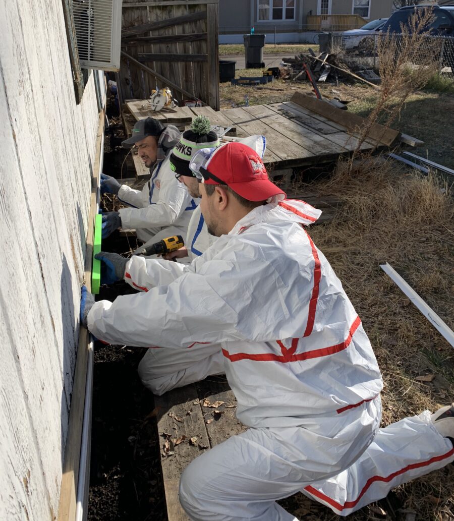 A group of men in hazmat suits repair the foundation of a trailer home.