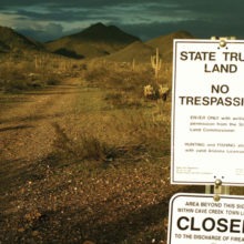 State Trust Land sign posted at road with rugged landscape. Cacti and sage are throughout the landscape.